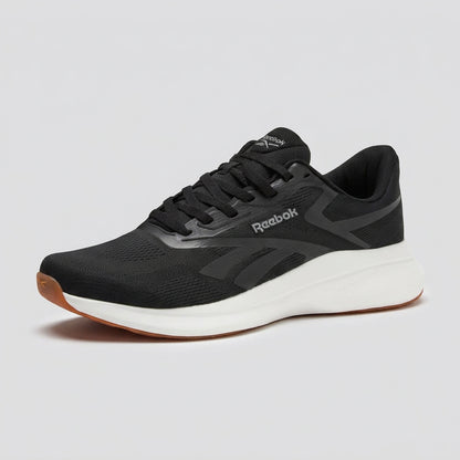 A Reebok athletic sneaker in black and dark gray with a thick white midsole and a gum brown outsole, shown from a three-quarters angle on a white background.