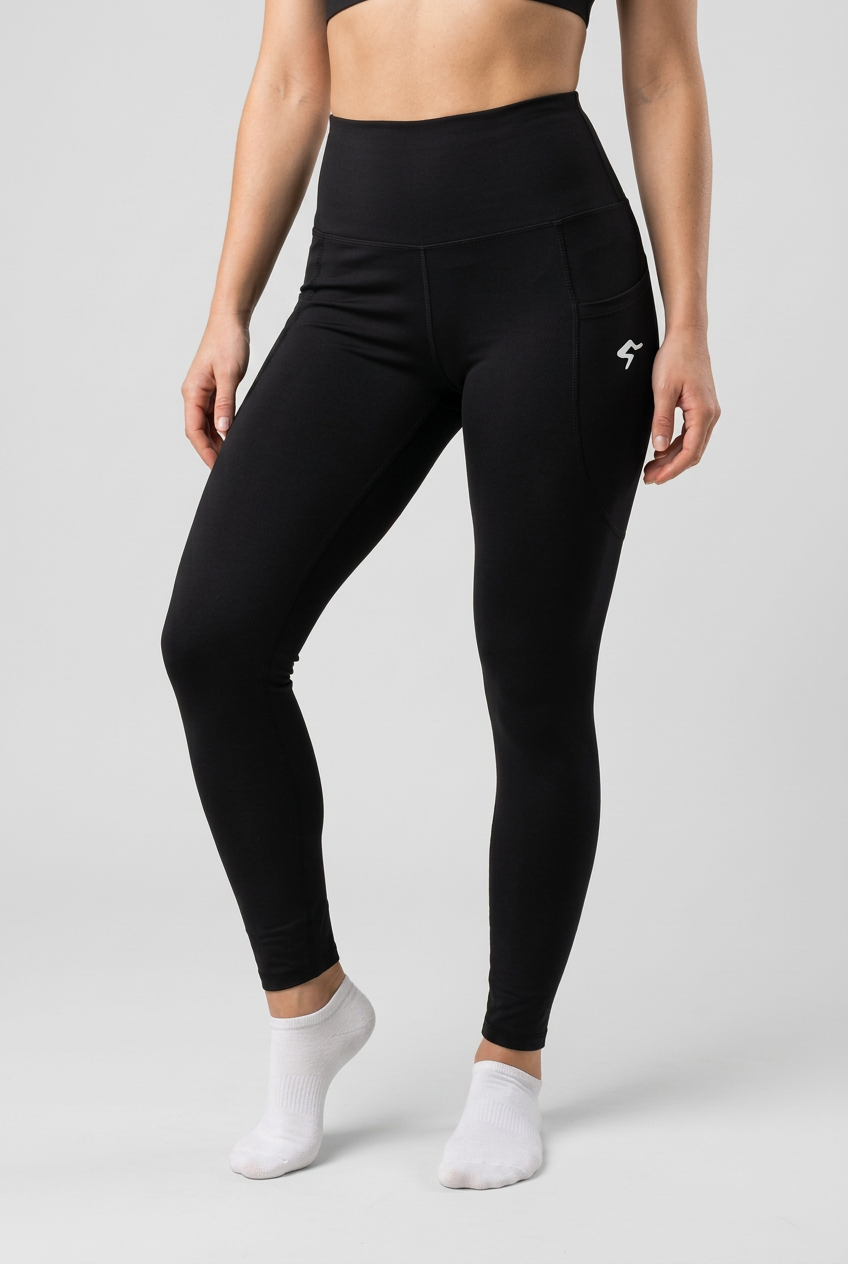 A model wearing black high-waisted athletic leggings with a small white logo on the upper right hip, paired with white ankle socks, against a solid light beige background.
