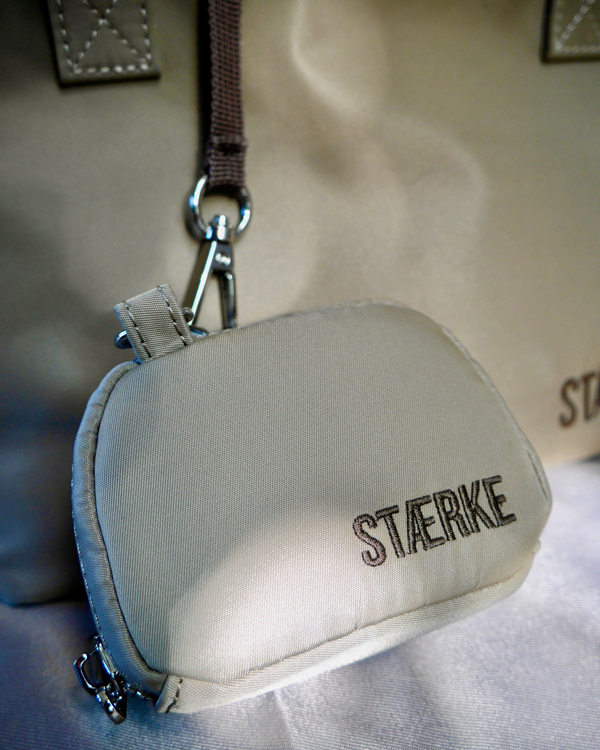 Sand-colored round zip jewelry pouch with silver keyring clip and gold logo text