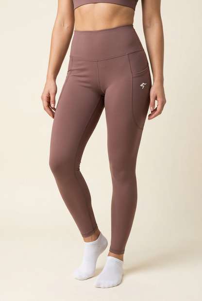 A model wearing high-waisted, form-fitting leggings in a dusty mauve-brown color, featuring a side cargo pocket with a small white logo. The model is also wearing white ankle socks and a matching sports bra against a solid beige background.