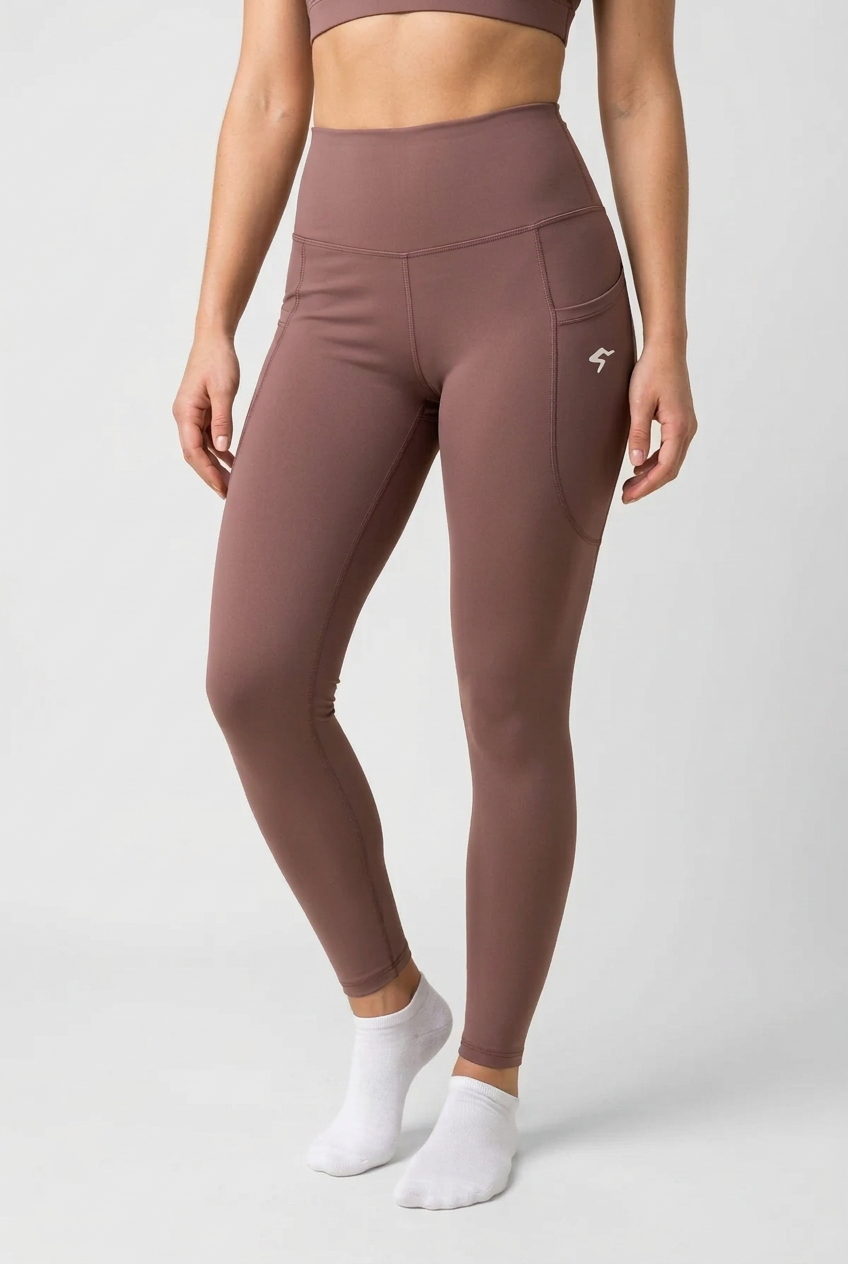 A model wearing high-waisted, form-fitting leggings in a dusty mauve-brown color, featuring a side pocket with a small white logo. The model is also wearing a matching sports bra and white ankle socks, photographed against a neutral light gray studio background.