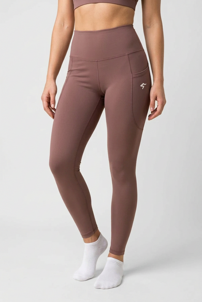 A model wearing high-waisted, form-fitting leggings in a dusty mauve-brown color, featuring a side pocket with a small white logo. The model is also wearing a matching sports bra and white ankle socks, photographed against a neutral light gray studio background.