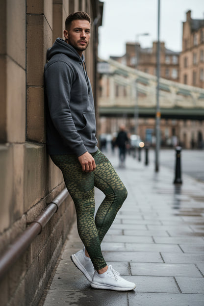 Man in gray hoodie and green patterned leggings leaning against a wall on a city street.