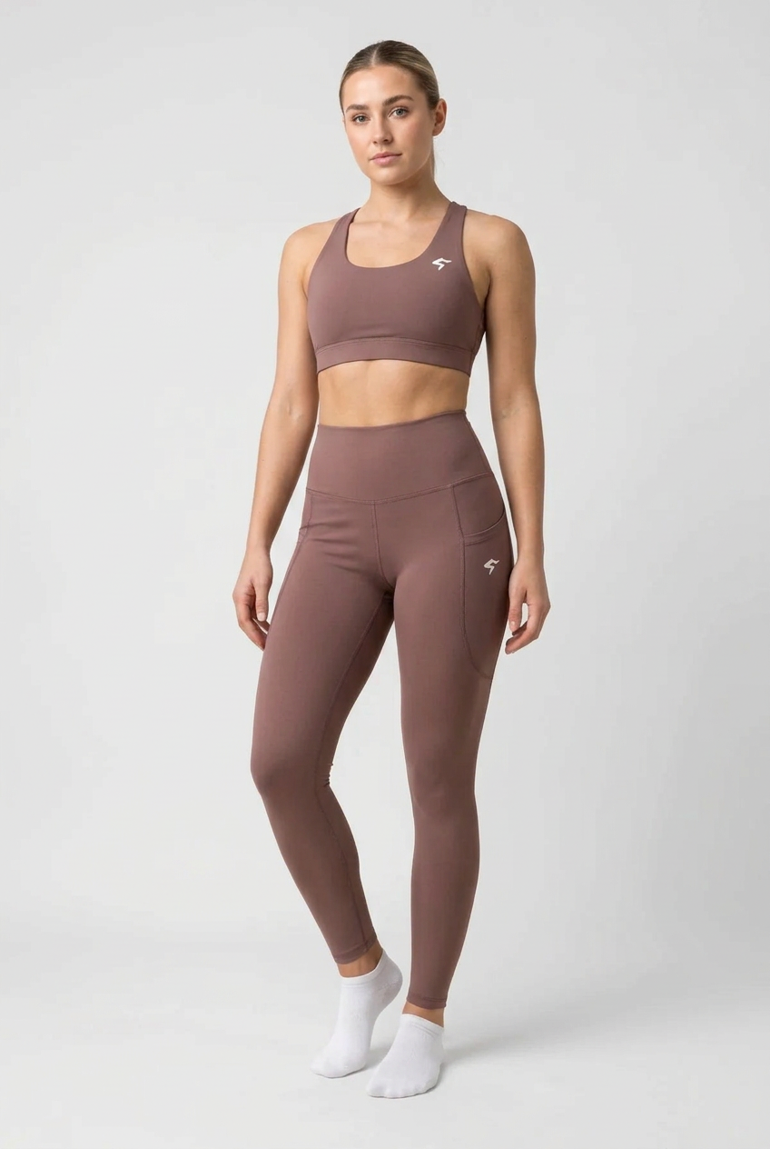 A woman modeling a matching athletic set consisting of a sports bra and high-waisted leggings in a dusty mauve-brown color, paired with white socks, against a light gray studio background.