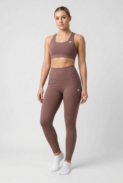 A woman modeling a matching athletic set consisting of a sports bra and high-waisted leggings in a dusty mauve-brown color, paired with white socks, against a light gray studio background.