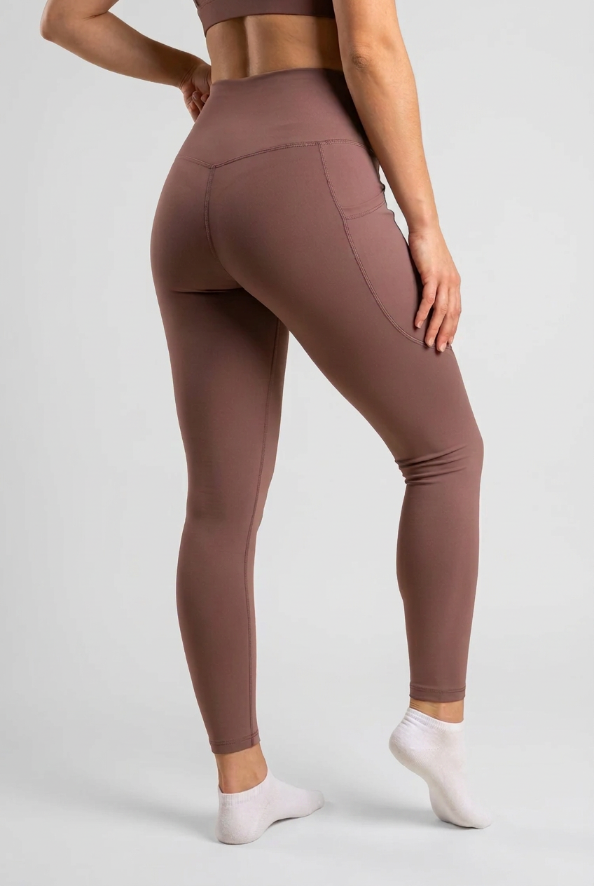 A model wearing high-waisted, form-fitting leggings in a dusty mauve-brown color, shown from a rear three-quarter angle against a solid cream background.