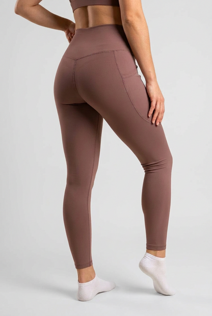 A model wearing high-waisted, form-fitting leggings in a dusty mauve-brown color, shown from a rear three-quarter angle against a solid cream background.