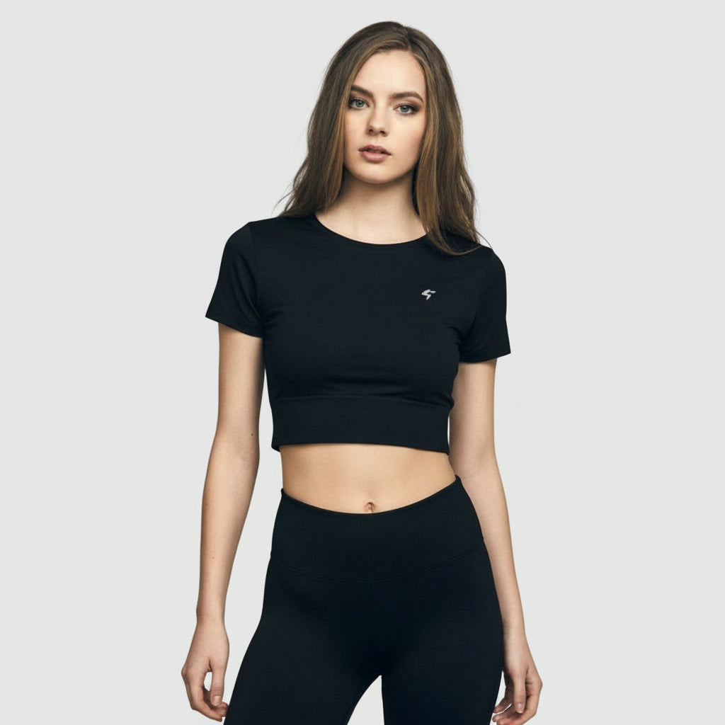 A woman wearing a black slim-fit crop tee with short sleeves, standing in front of a brick wall. The tee appears to be made of a soft and durable fabric.