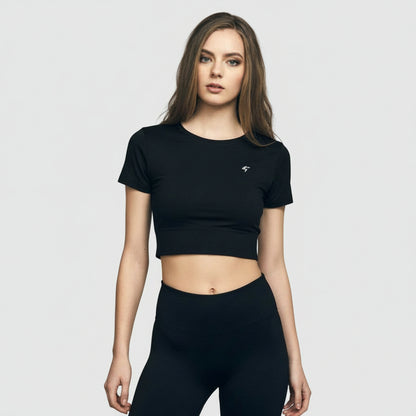 A woman wearing a black slim-fit crop tee with short sleeves, standing in front of a brick wall. The tee appears to be made of a soft and durable fabric.