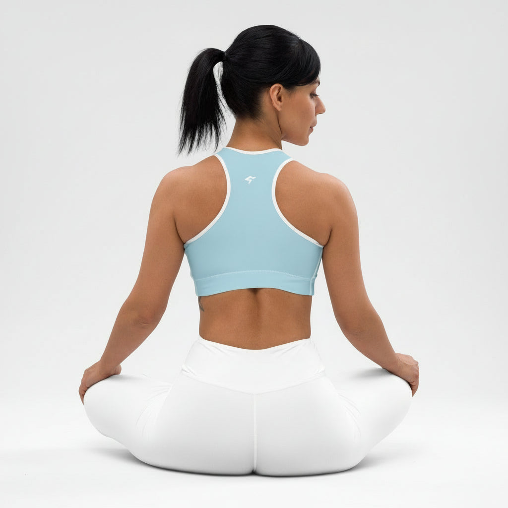 Woman sitting on a white yoga cushion wearing a light blue sports bra and white pants against a plain background