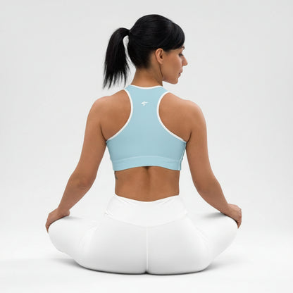 Woman sitting on a white yoga cushion wearing a light blue sports bra and white pants against a plain background