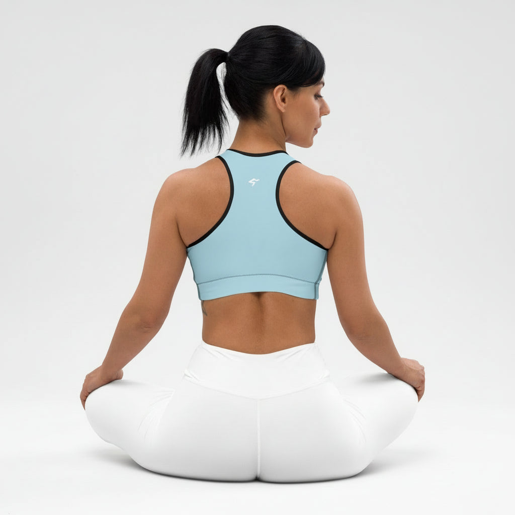 Woman sitting on a white yoga cushion wearing a light blue sports bra and white pants against a plain background