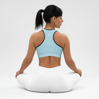 Woman sitting on a white yoga cushion wearing a light blue sports bra and white pants against a plain background
