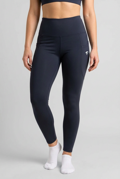 A model wearing high-waisted navy blue athletic leggings with a small white logo on the left hip, paired with white ankle socks, against a solid beige background.