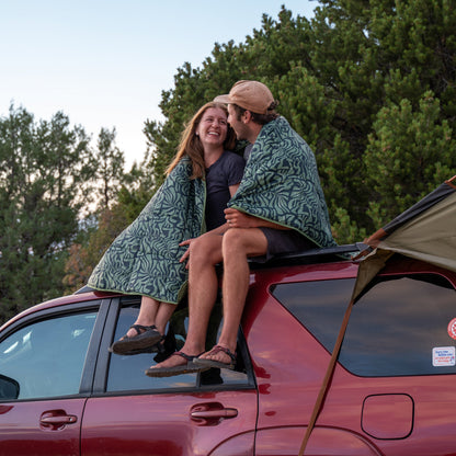 Camping blanket with teal leaf pattern draped across two people sitting on a car